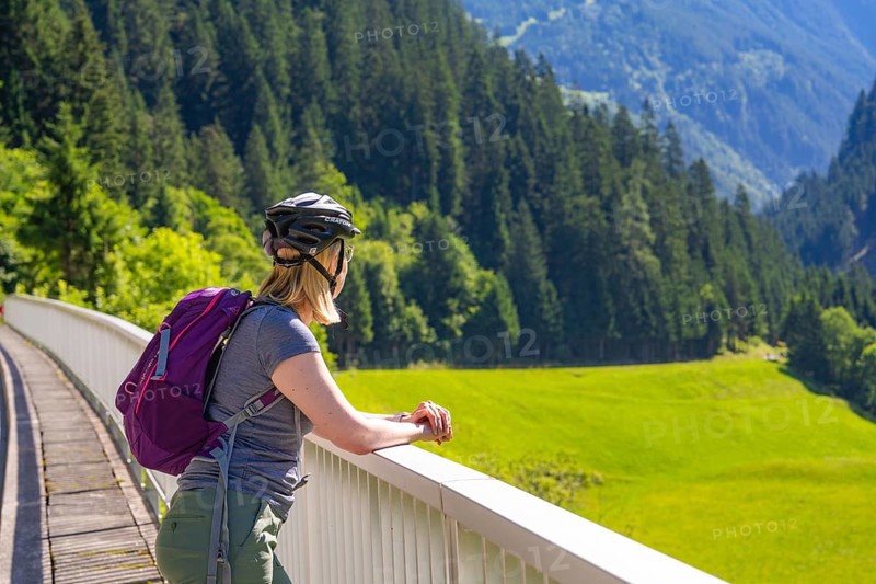 A woman with a bicycle helmet and a purple rucksack leans on a balcony and looks at a green hilly landscape, Zillertal, Austria, Germany, Europe