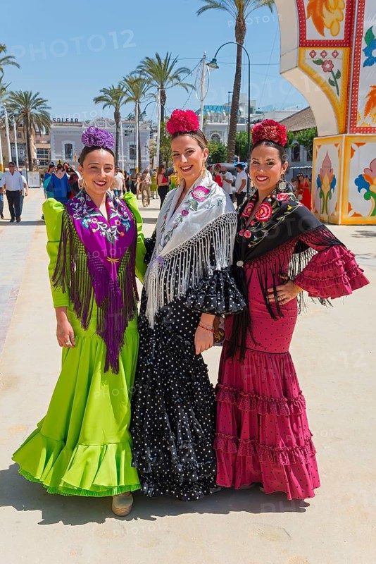 Three woman in colourful flamenco dresses, with flowers in their hair, pose on a sunny street with palm trees and festive background, Feria de la Manzanilla, Feria de Sanlúcar, Sanlúcar de Barrameda, Sanlucar, Cádiz, Cadiz, Andalusia, Spain, Europe