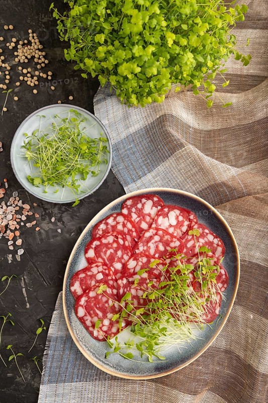 Slices of smoked cervelat salami sausage with spinach microgreen, salt and pepper on black concrete background and beige textile. Top view, flat lay, close up