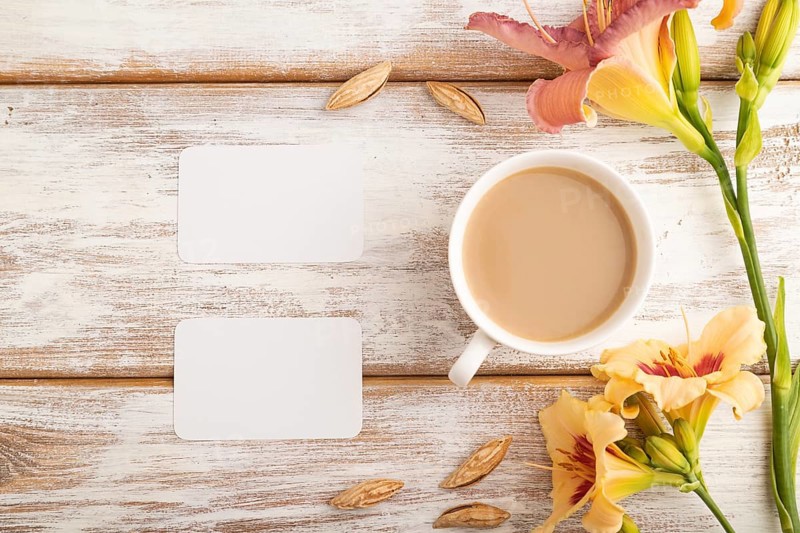 White paper business card mockup with orange day-lily flower and cup of coffee on white wooden background. Blank, top view, flat lay, copy space, still life. spring concept