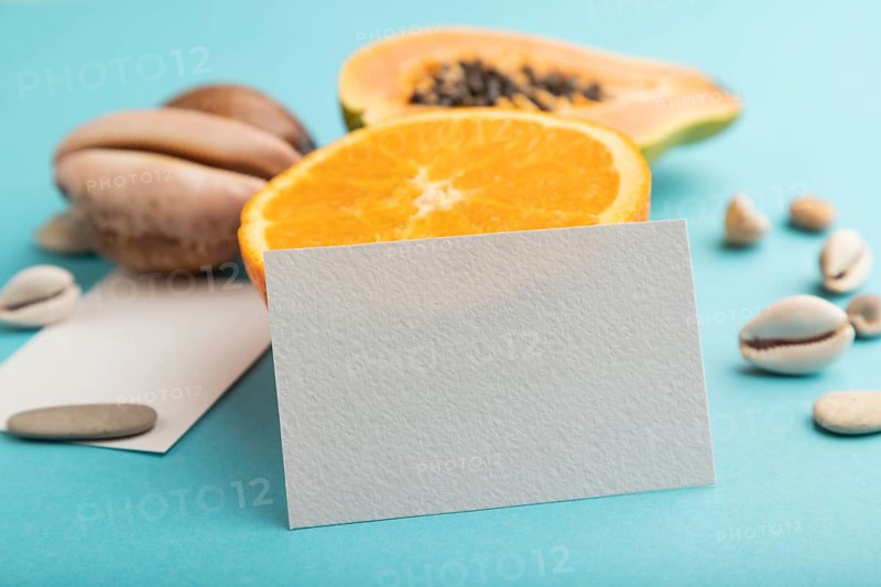White paper business card with ripe cut papaya, orange, seashells on blue pastel pastel background. Side view, copy space. Tropical, healthy food, vacation, holidays concept