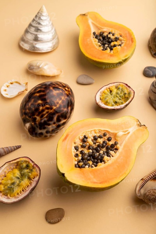 Ripe cut papaya, passion fruit, seashells, pebbles on orange pastel background. Side view, close up. Tropical, healthy food, vacation, holidays concept