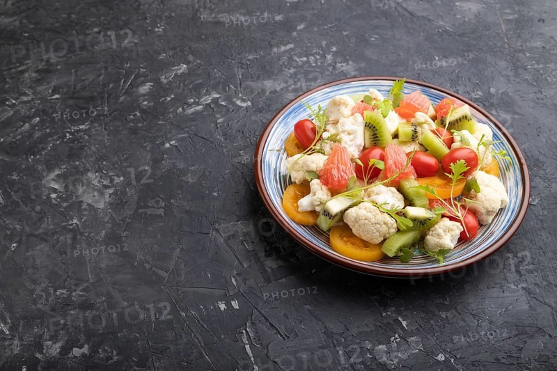 Vegetarian salad of cauliflower cabbage, kiwi, tomatoes, microgreen sprouts on black concrete background. Side view, close up, copy space