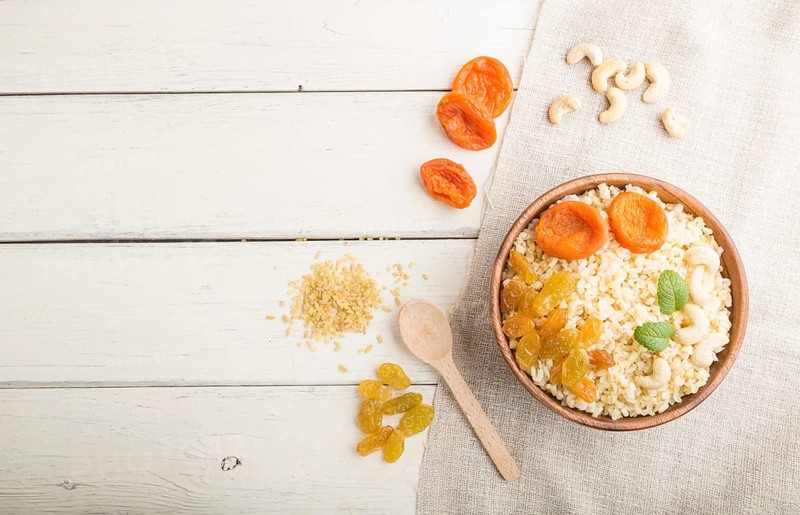 Bulgur porridge with dried apricots, raisins and cashew in wooden bowl on a white wooden background and linen textile. Top view, flat lay, copy space. Turkish traditional cuisine