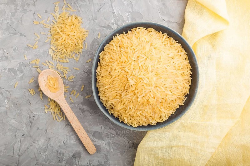 Blue ceramic bowl with raw golden rice and wooden spoon on a gray concrete background and yellow textile. Top view, flat lay, close up