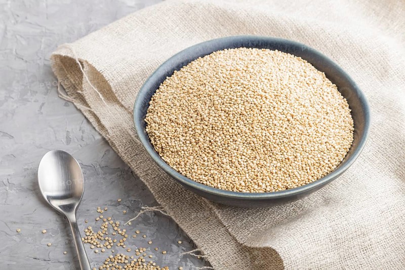 Blue ceramic bowl with raw white quinoa seeds on a gray concrete background and linen textile. Side view, close up