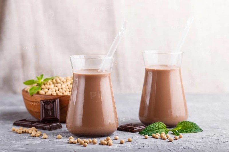 Organic non dairy soy chocolate milk in glass and wooden plate with soybeans on a gray concrete background. Vegan healthy food concept, close up, side view