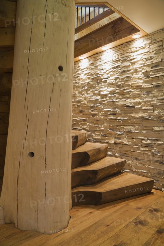 Twisting wooden staircase with brown stained half log steps attached to large bleached supporting central tree trunk post plus illuminated lights shining on tan natural stone wall in basement leading to ground floor inside handcrafted Eastern white pine Scandinavian log cabin home, Quebec, Canada, North America