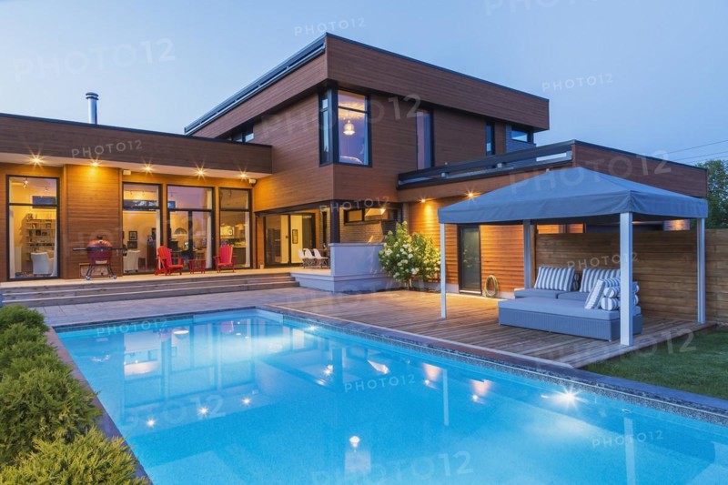 Rear of modern cube style home with stained horizontal wood cladding, in-ground swimming pool and wooden deck with gazebo canopy at dusk, Quebec, Canada, North America