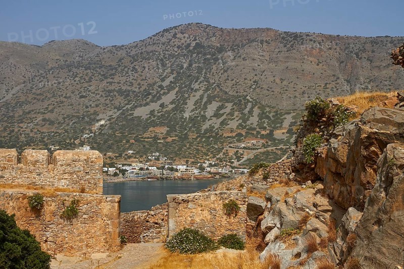 The remains of ancient stone structures in front of a mountainous landscape with a nearby village, Venetian Sea Fortress, Leper Island, Spinalonga, Elounda Bay, Mirabello Bay, East Crete, Crete, Greek Islands, Greece, Europe