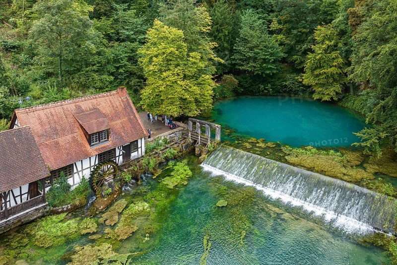 Blautopf Blaubeuren with industrial monument Hammerschmiede, source of the little river Blau in a landscape with forest. Karst spring, geotope and geopoint of the UNESCO Swabian Alb Geopark, tourist attraction. The popular excursion destination is now being thoroughly renovated and will therefore be closed to visitors until the end of 2028. Drone photo. Blaubeuren, Baden-Württemberg, Germany, Europe