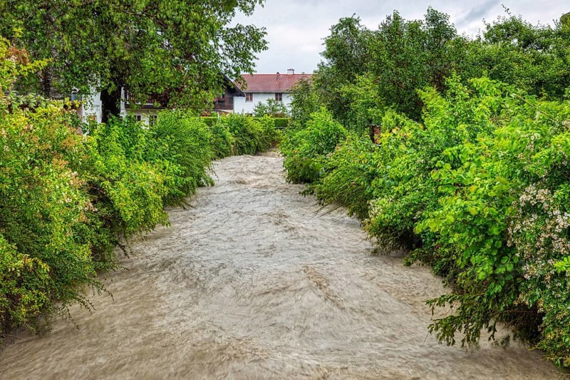 A heavily flooded river carves its way through a densely overgrown green landscape with houses, Aubach, Au