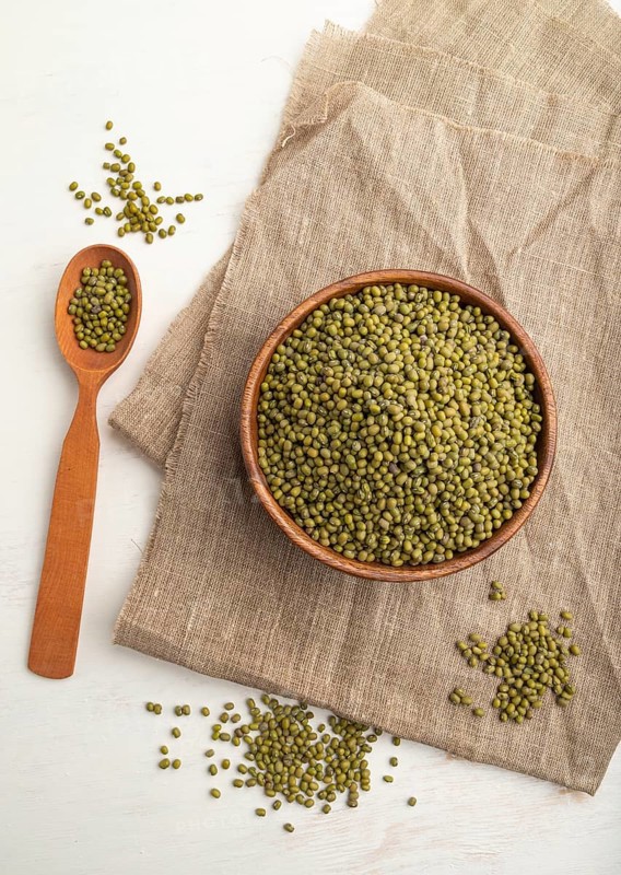 Wooden bowl with raw green mung bean and wooden spoon on a white background and linen textile. Top view, flat lay, close up
