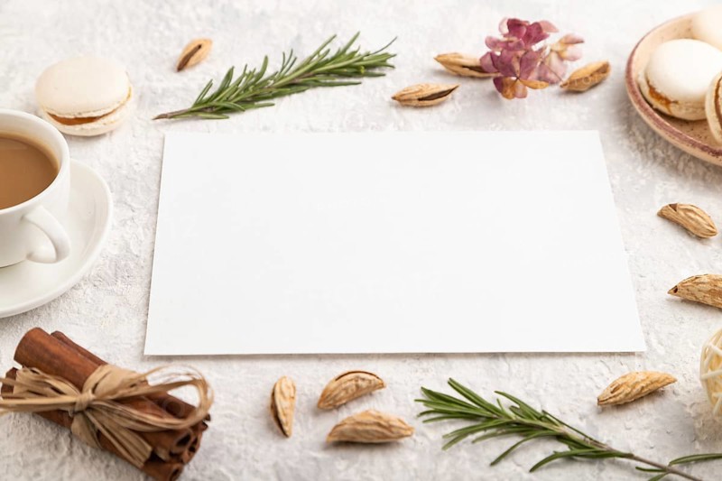 White paper sheet mockup with cup of coffee, almonds and macaroons on gray concrete background. Blank, business card, side view, still life