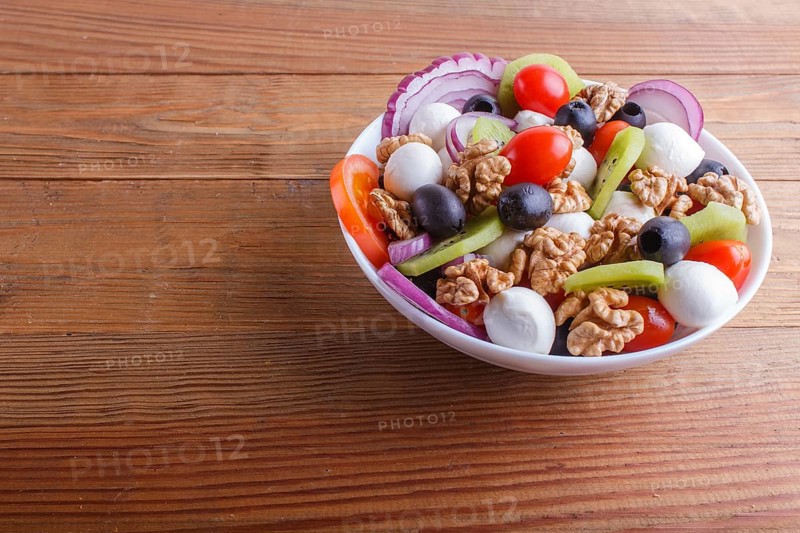 Salad with fresh cherry tomatoes, mozzarella cheese, olives, kiwi, onion and walnuts on brown wooden background, copy space, close up