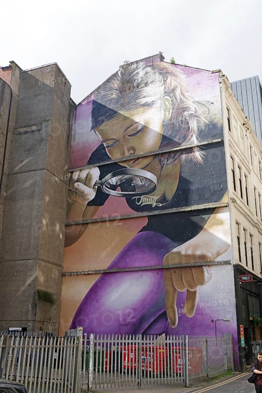 A mural shows a woman looking at something with a magnifying glass. In the background are, Mural Trail, Glasgow, Scotland, Great Britain