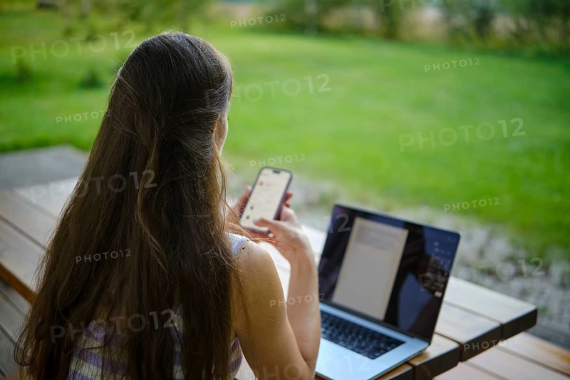 A multitasking woman using a laptop and smartphone in a serene garden setting while working on wooden terrace in the evening