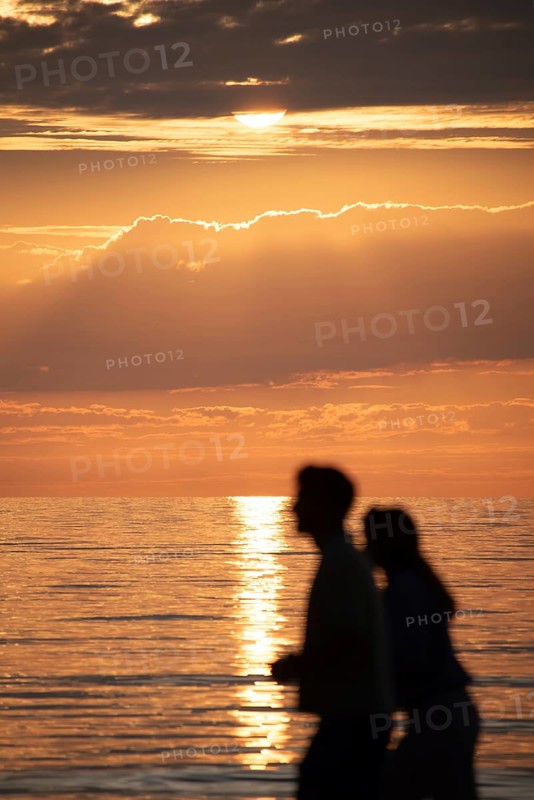 Two people walking along the sea at sunset, their silhouettes against a bright sky and calm water, Dierhagen, Mecklenburg-Vorpommern, Germany, Europe