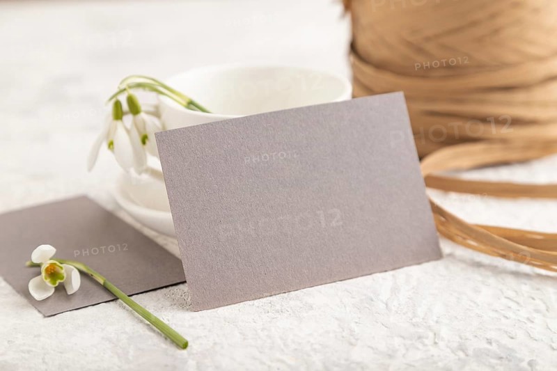 Gray paper invitation card, mockup with galanthus snowdrop flowers on gray concrete background. Blank, side view, still life, copy space, wedding invitation