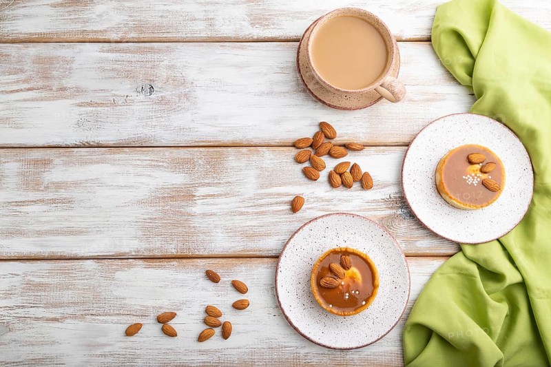 Sweet tartlets with almonds and caramel cream with cup of coffee on a white wooden background and green textile. top view, flat lay, copy space