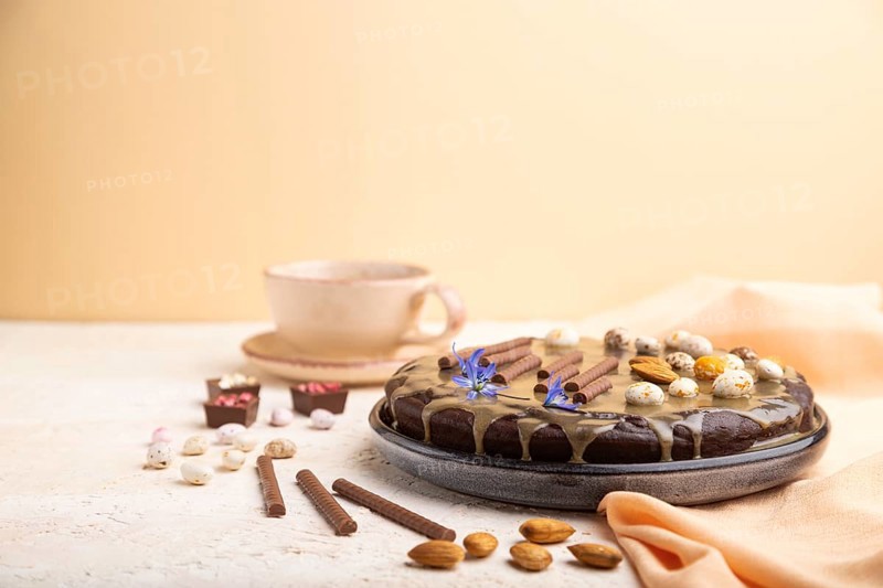 Homemade chocolate brownie cake with caramel cream and almonds with cup of coffee on a white and orange background and orange textile. Side view, copy space, selective focus