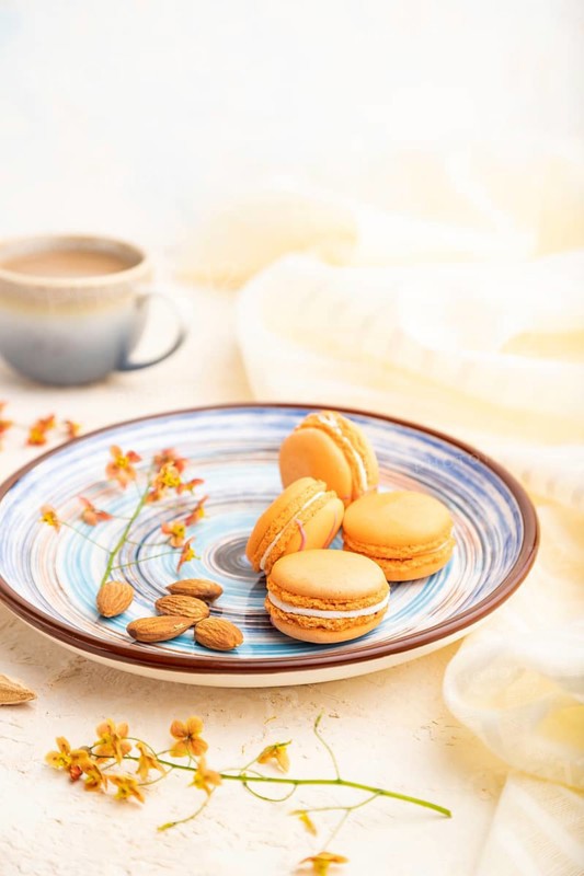 Orange macarons or macaroons cakes with cup of coffee on a white concrete background and linen textile. Side view, close up, selective focus
