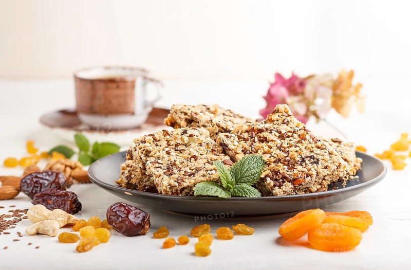 Homemade granola from oat flakes, dates, dried apricots, raisins, nuts in blue ceramic plate with a cup of coffee on a white wooden background. Side view, close up, selective focus