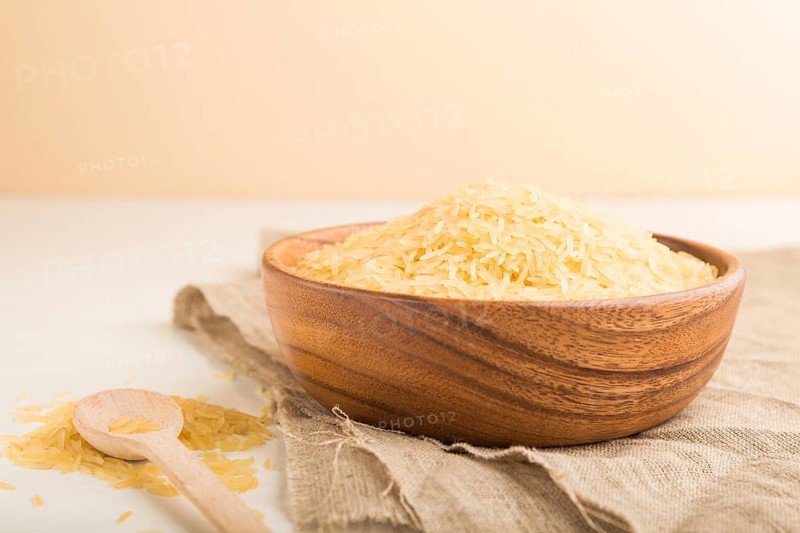 Wooden bowl with raw golden rice and wooden spoon on a white wooden background and linen textile. Side view, close up, selective focus
