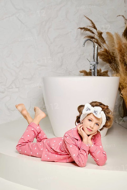 Smiling young girl laying down relaxed on the floor in the bathroom, wearing a cozy pink pajama and hair band