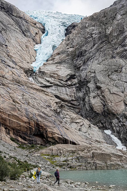 Hikers marvel at the melting Kjenndalsbreen glacier and a mountain lake surrounded by steep rock faces in Norway Europe