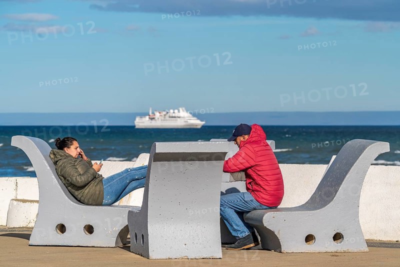 People in warm winter clothing sit on benches on the banks of the Strait of Magellan, city of Punta Arenas, Patagonia, Chile, South America