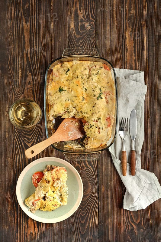 A baked casserole showcasing layers of cannelloni pasta, vegetables and melted cheese accompanied by a glass of wine and neatly arranged utensils
