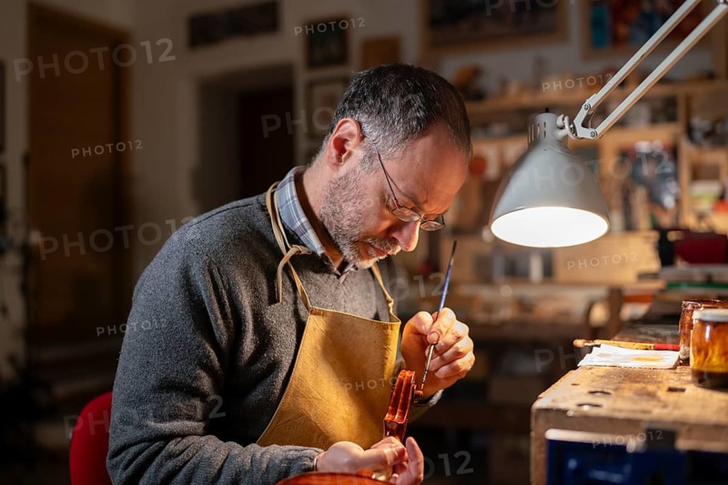 Violin maker working on a violin in his workshop, using a small brush and varnish, illuminated by a lamp, surrounded by tools and materials