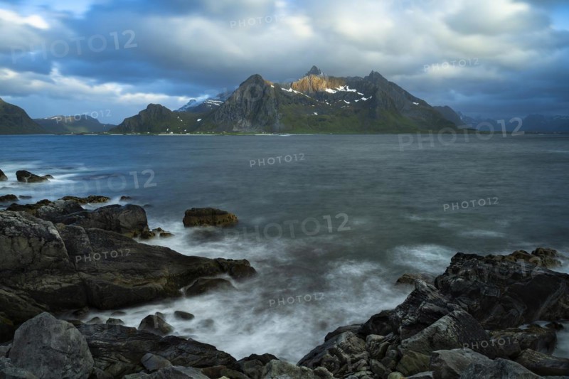 Landscape with sea and mountains on the Lofoten Islands, view across the fjord to the small town of Flakstad and the mountain Flakstadtinden as well as other mountains. Rocks in the foreground. At night at the time of the midnight sun, some clouds in the sky. Long exposure, blurred clouds, sun on a mountain peak. Early summer. Flakstadoya, Lofoten, Norway, Europe