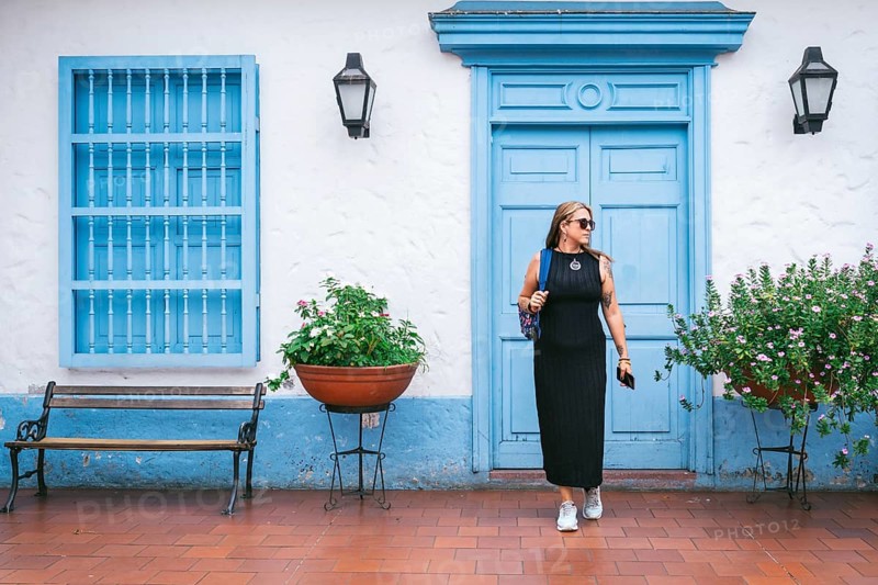 A fashionable woman in a black dress and sneakers walks past a blue colonial-style door and window adorned with flowers. The vibrant scene blends traditional architecture with modern fashion.