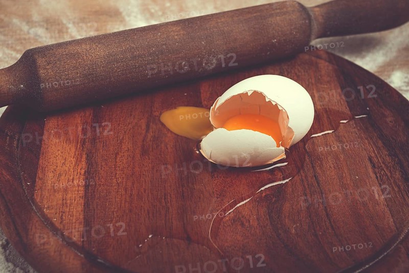 A broken egg with its yolk spilling onto a wooden cutting board sits beside a rolling pin, indicating the start of cooking or baking in a home kitchen.