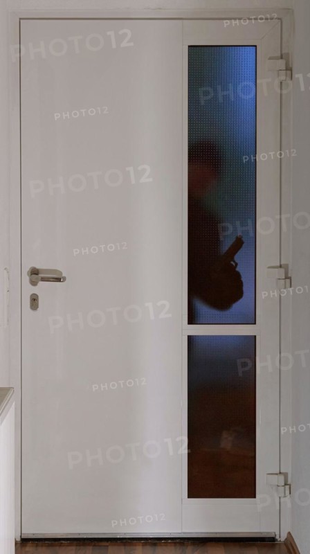 A burglar stands in front of an entrance door in the dark with a gun and looks through the glass pane into the flat or house