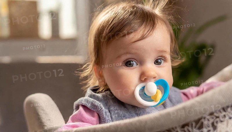 A baby with a blue dummy and blond hair sits in a cot and looks attentively, AI generated, AI generated