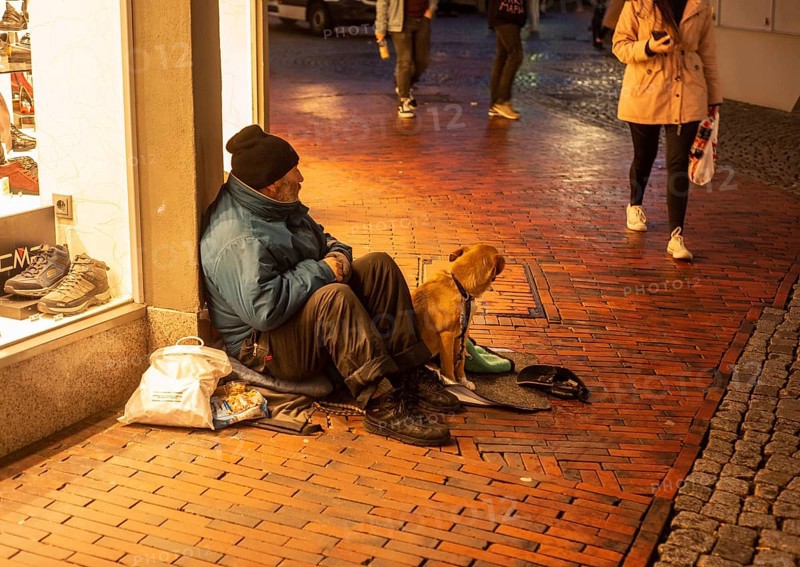 Homeless man with a dog sits in a shopping mall as people walk by in Husum, Schleswig-Holstein, Germany, Europe