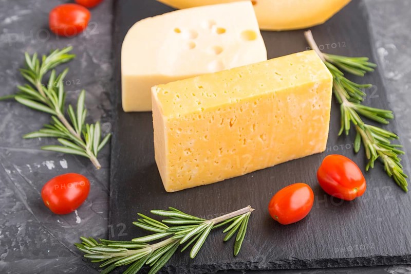 Various types of cheese with rosemary and tomatoes on black slate board on a black concrete background. Side view, close up, selective focus