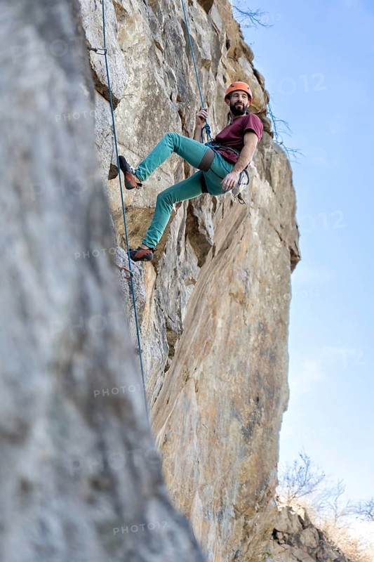 A young man climbs a challenging cliff route, secured by a rope, showcasing strength and determination against the rugged rock face under a clear sky, capturing the thrill and intensity of the ascent