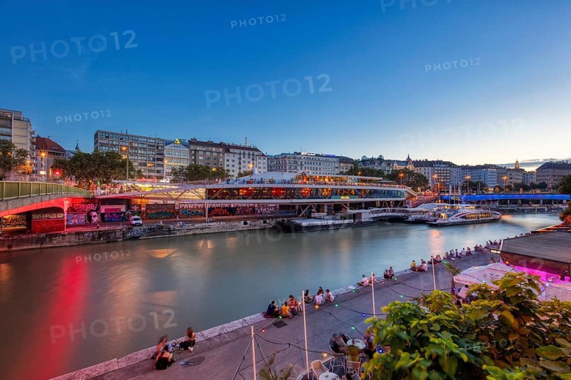 Evening scene by the river with illuminated promenade and people on the riverbank, relaxed atmosphere, Vienna