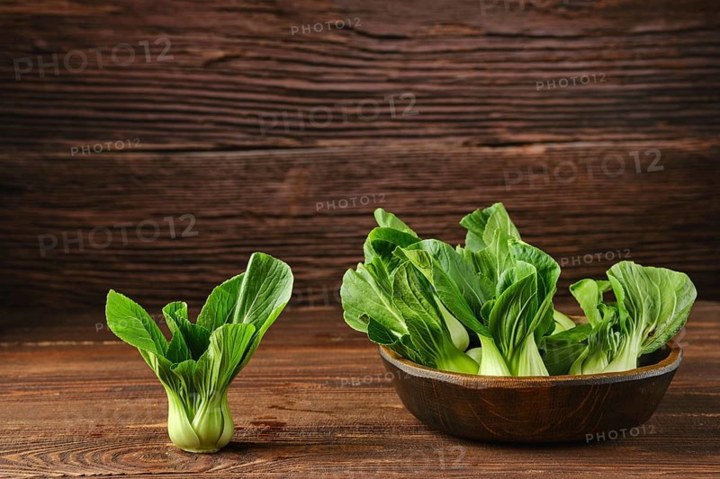 A collection of vibrant bok choy in a rustic wooden bowl on a wooden kitchen table, with one smaller bok choy beside it, highlighting their fresh green color and healthy appearance.