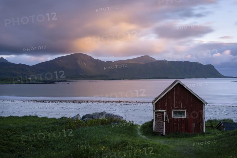 A red wooden hut on the sandy beach of Ramberg (Rambergstranda), with the sea behind it. At night at the time of the midnight sun. Clouds in the sky, the sun shines on clouds and mountains. Early summer. Long exposure, blurred clouds. Ramberg, Flakstadoya, Lofoten, Norway, Europe