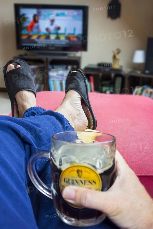Couch potato, lazy man in comfy chair with pint of beer wearing worn slippers with big toes sticking through and watching television in living room