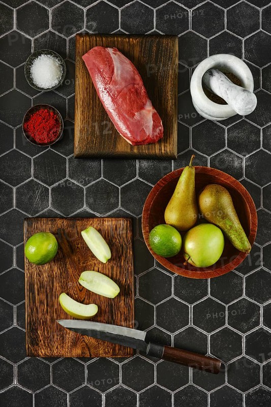 A variety of fresh ingredients lie on chopping wooden boards, including raw meat, vibrant apples and pears ready to be transformed into a gourmet dish