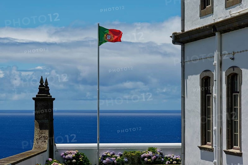 Portuguese flag flying next to a white building with a view of the blue sea and blooming flowers, Farol da Ferraria lighthouse, Sao Miguel Island, Azores, Portugal, Europe