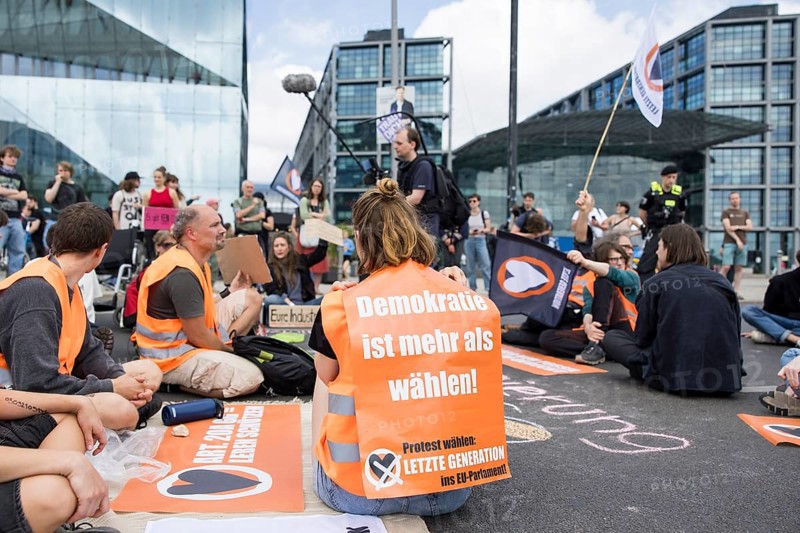 Last Generation activist with poster Democracy is more than voting! for the upcoming European elections at the occupation of Rahel-Deer-Straße in front of Berlin Central Station, Berlin, 25 May 2024