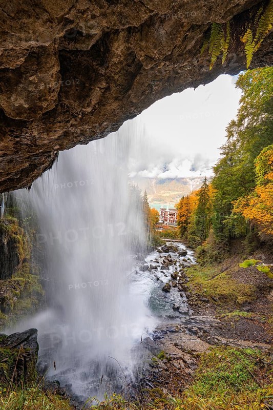 A waterfall flows through a forest with a view of a distant building, Lake Brienz, Giessbach Waterfall, Switzerland, Europe
