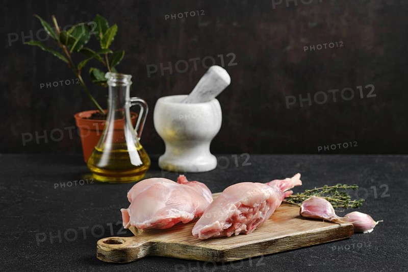 Fresh rabbit legs on a wooden board, accompanied by herbs, garlic, olive oil, and a mortar and pestle in a rustic kitchen setting, perfect for meal preparation.
